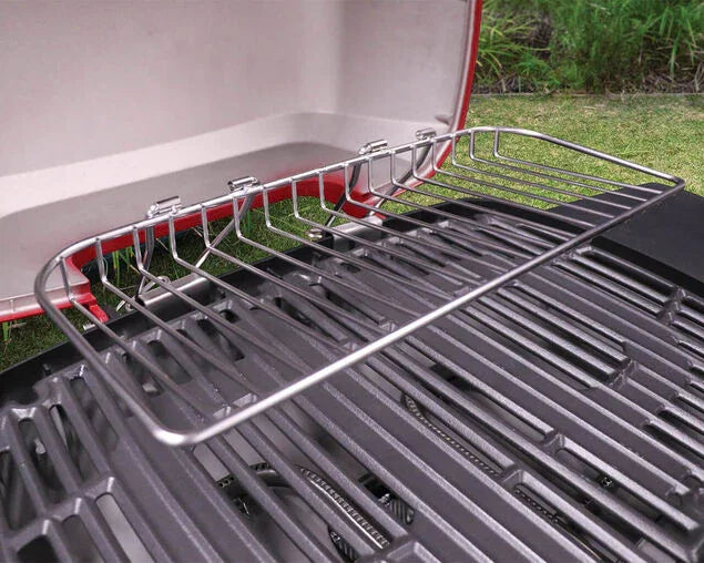 Stainless steel warming rack mounted on outdoor gas grill with black grates, garden background