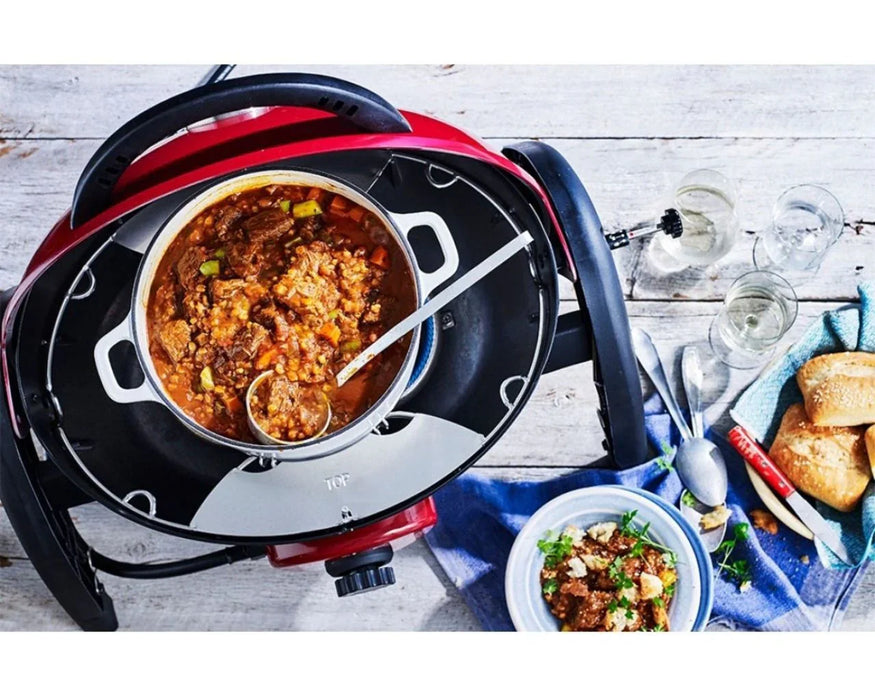 Portable outdoor grill with brown trivet kit, stew pot, bread, and serving utensils on table.