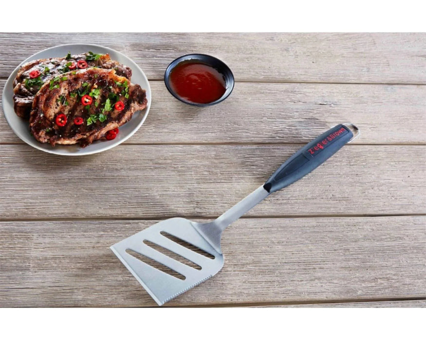 Stainless steel BBQ spatula with black handle on wooden table beside grilled steak and sauce.