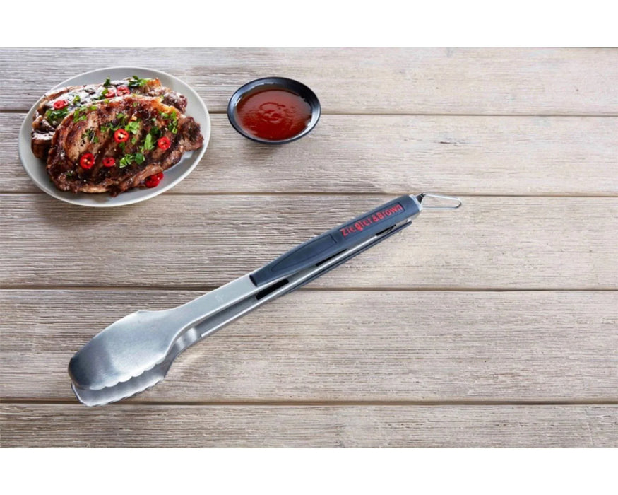 Stainless steel kitchen tongs beside grilled meat and sauce on wooden table.