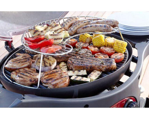 Outdoor grill with two-tier warming rack cooking assorted meats, sausages, and vegetables