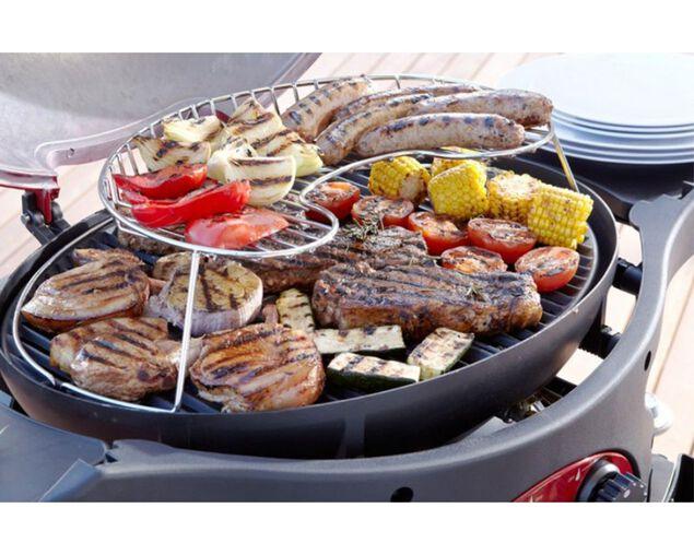 Outdoor grill with two-tier warming rack cooking assorted meats, sausages, and vegetables